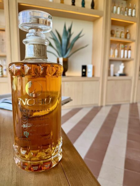 Masterclass au-delà de l'agave au Nobu Hotel Los Cabos A bottle of Nobu rare añejo tequila on a wooden table, with a plant and shelves of tequila in the background.