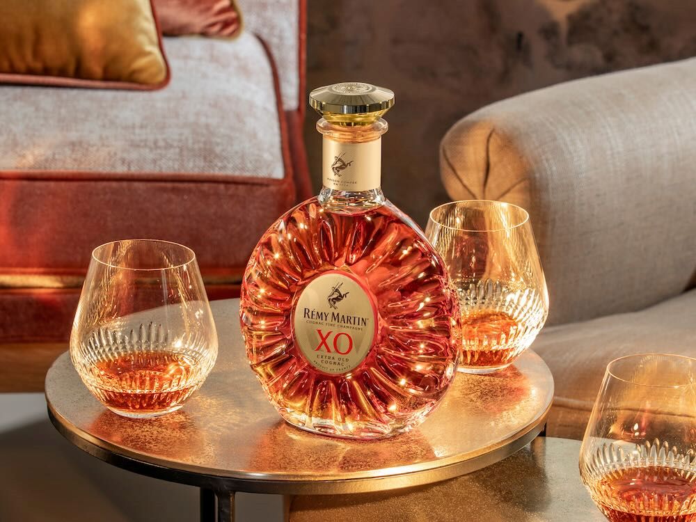 La bouteille Rémy Martin XO repose sur une table entourée de deux verres remplis de cognac, installés dans un salon élégant.