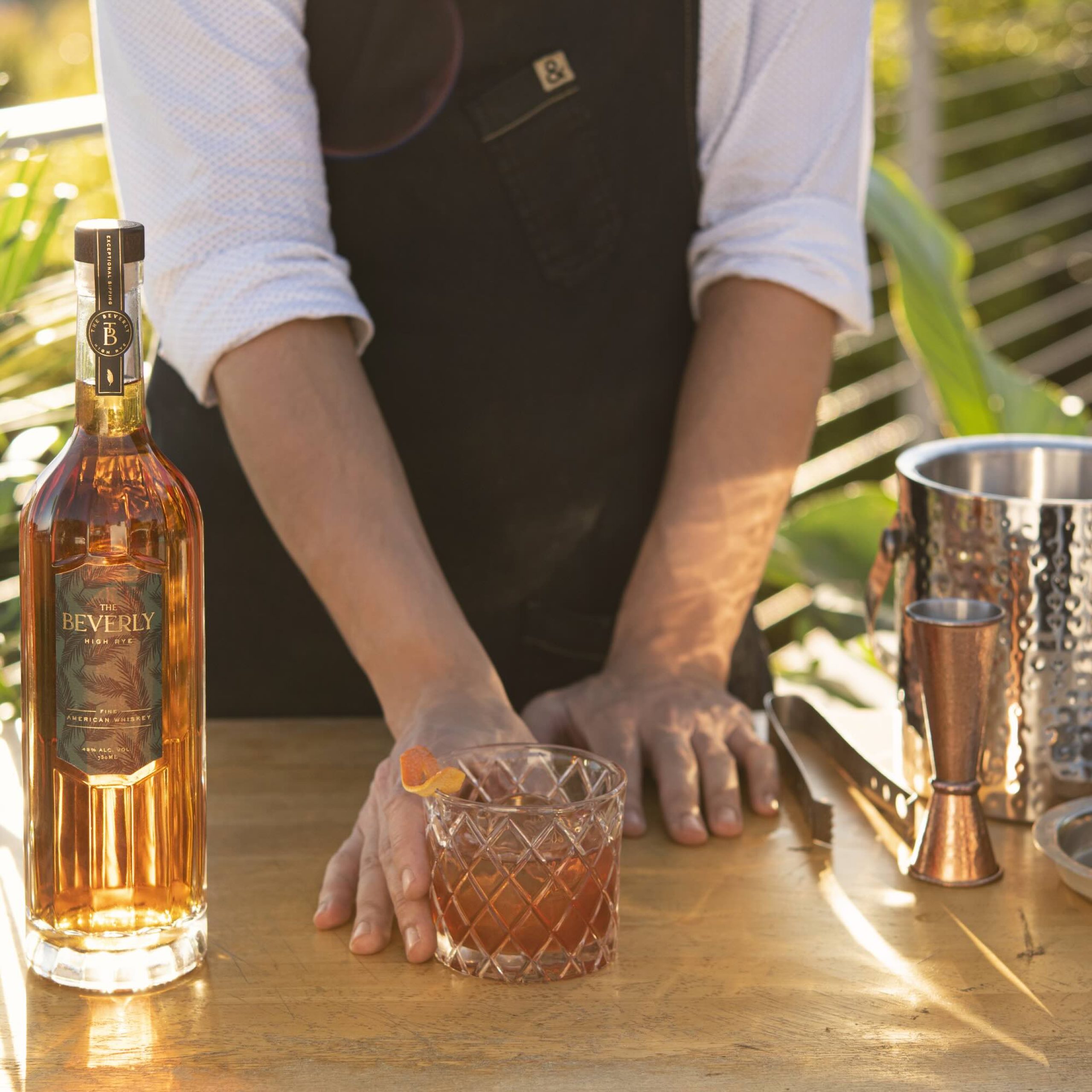 Bouteille de whisky Beverly High Rye à côté d'un verre à cocktail sur une table en bois, entourée de verdure et d'outils de barman.