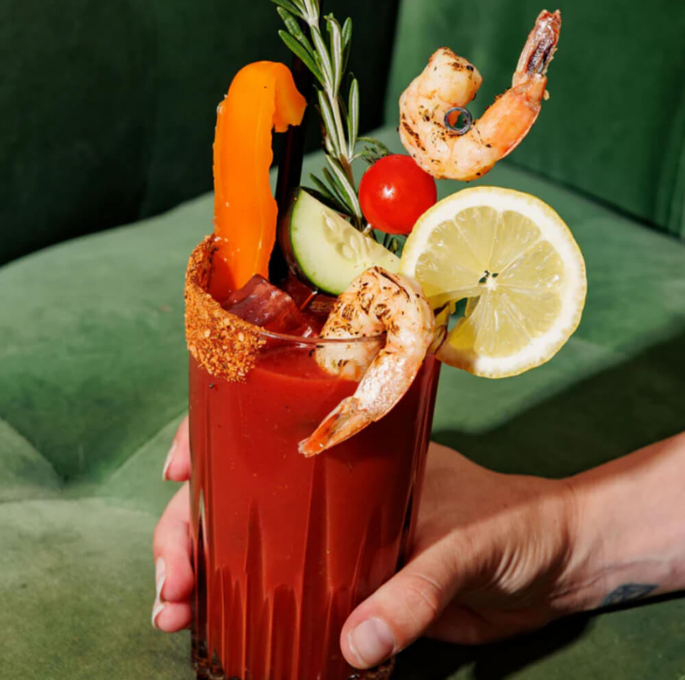 Une main tient un verre Bloody Mary garni de crevettes, de citron, de concombre, de tomate et d'une tranche de poivre sur fond vert.