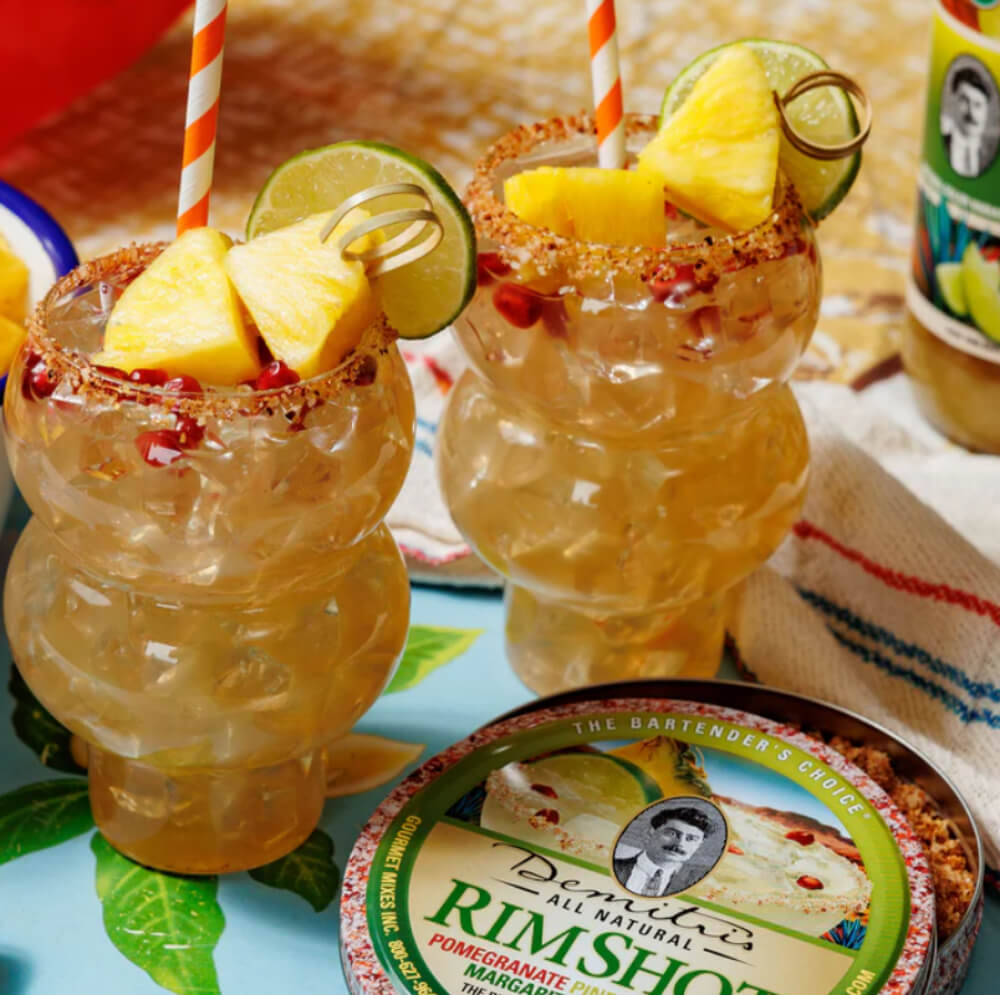 Deux cocktails tropicaux avec des garnitures d'ananas et de citron vert, servis dans une verrerie unique, à côté d'un récipient de sel aromatisé.