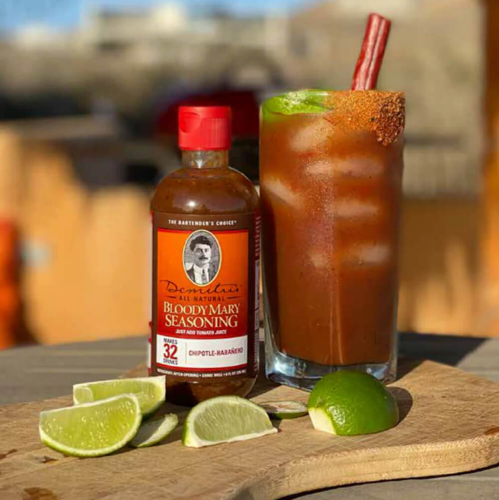 Bouteille d'assaisonnement Bloody Mary à côté d'un verre rempli d'un cocktail Bloody Mary, garni de citron vert et d'un bâton de céleri sur une surface en bois.