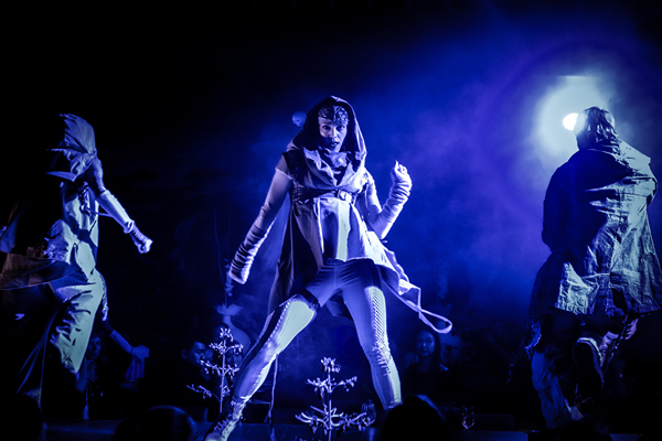 Trois artistes vêtus de costumes dramatiques se lancent dans une danse théâtrale sous un éclairage bleu, créant une expérience culinaire immersive.