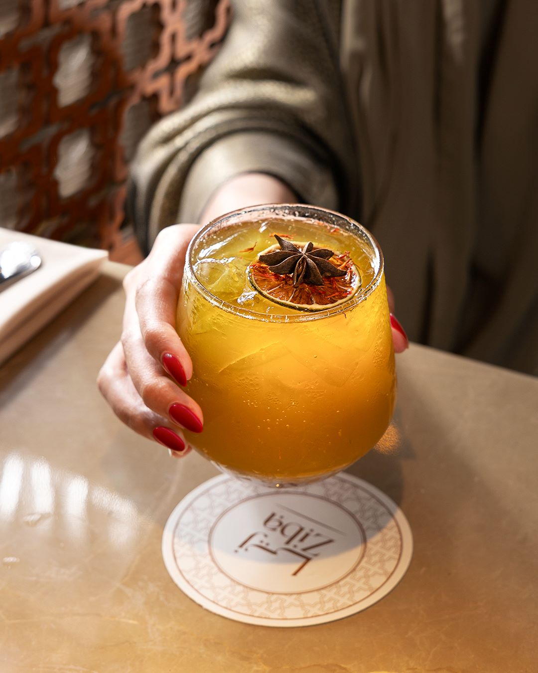 Une personne tenant un verre de boisson jaune garni d'anis étoilé et d'une tranche d'agrumes séchés sur un dessous de verre.