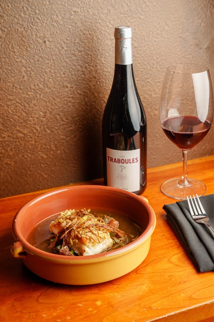 Un bol de poisson assaisonné garni d'échalotes croustillantes, une bouteille de vin et un verre de vin rouge sur une table en bois.