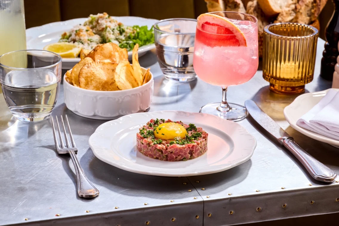 Une assiette de tartare de bœuf garnie d'un jaune d'œuf, accompagnée de chips croustillantes et d'un cocktail rose sur une table à manger.
