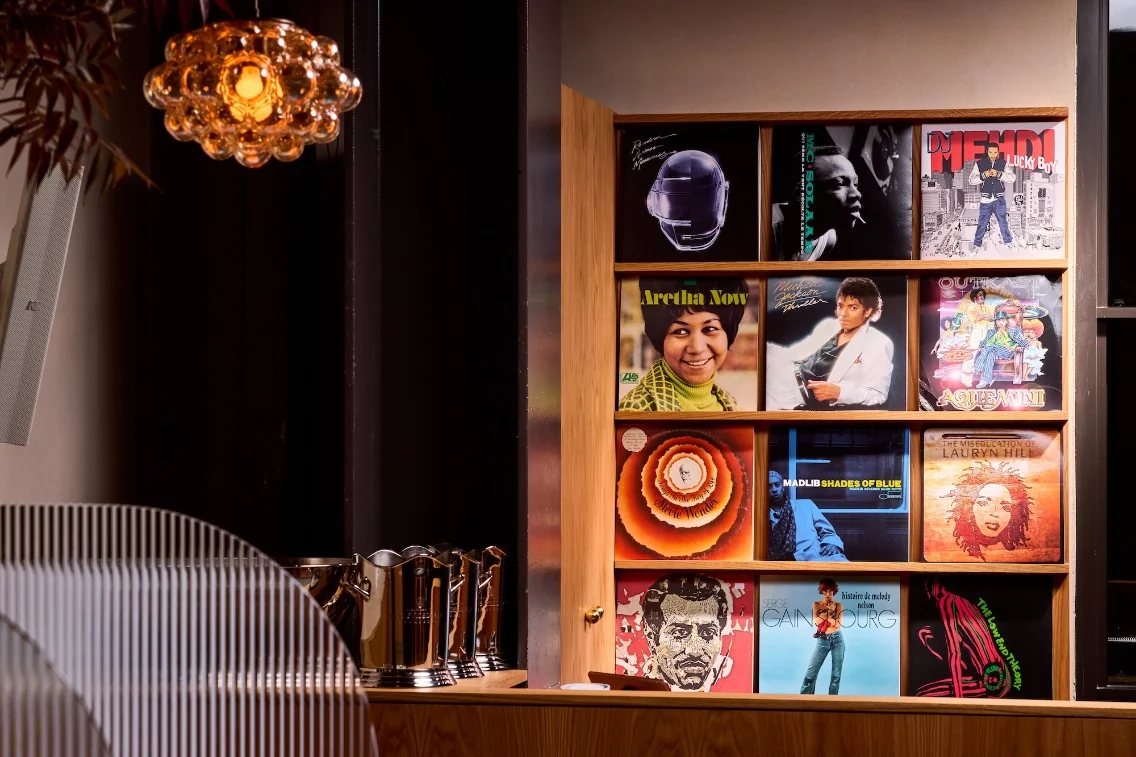 Une exposition de disques vinyles sur une étagère en bois, mettant en vedette divers artistes emblématiques et un luminaire décoratif au-dessus.