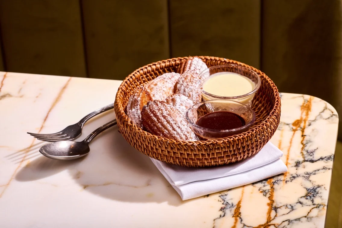 Un panier de madeleines saupoudré de sucre en poudre, accompagné de bols de sauces chocolat et vanille.