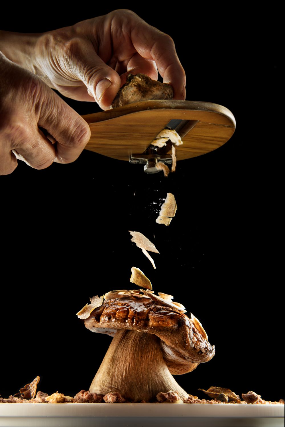 Mains râpant une truffe sur un cèpe, avec des copeaux tombant sur le plat sur fond noir.