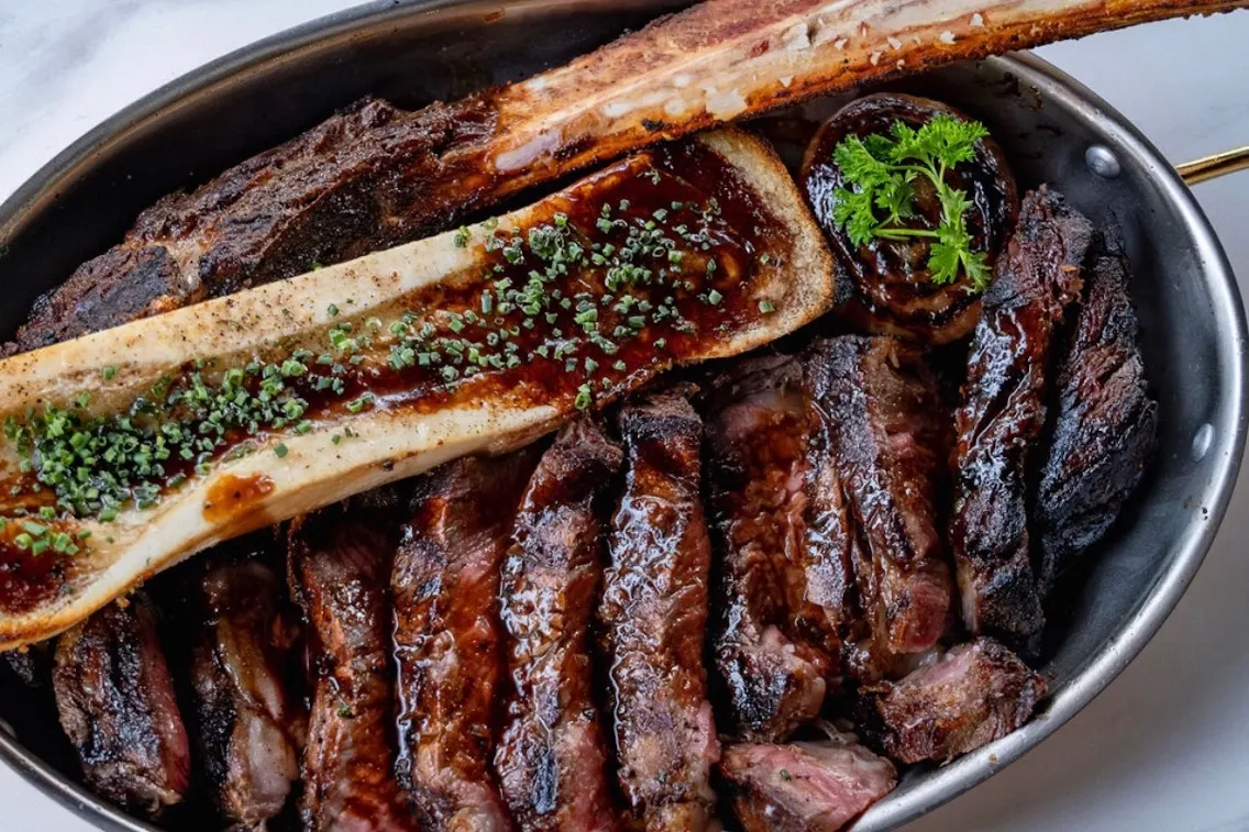 Un plateau de viandes grillées comprenant une côte de bœuf, de l'os à moelle et garni d'herbes, servi dans un plat rustique en métal.