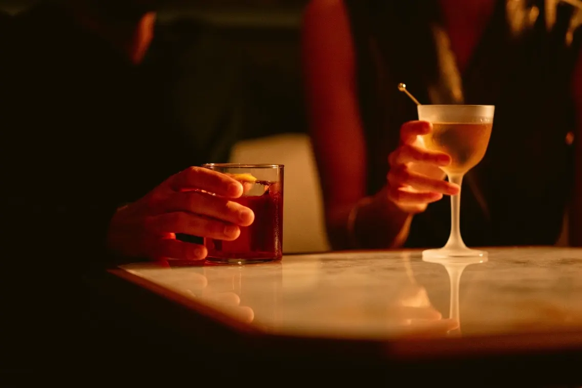 Deux mains tenant des cocktails, l'une dans un verre avec un zeste de citron et l'autre dans un verre à pied, sur une table en marbre.